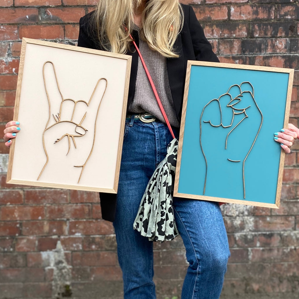 two hand painted wooden gesture signs. one cream with colour and Sign of the horns gesture. the other a blue background with a fist gesture.