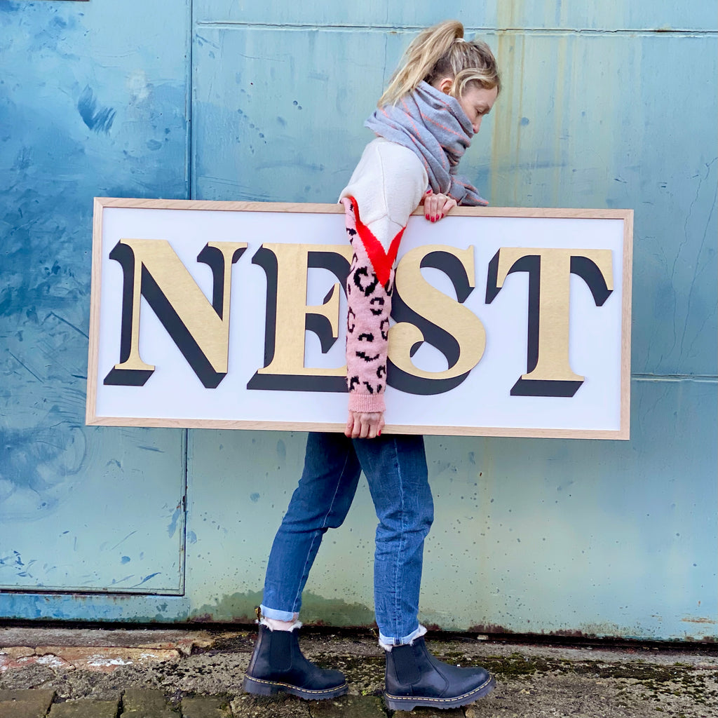 hand painted wooden layered one word sign with the word nest in gold and using a fancy font and a black drop shadow. the backdrop is a lavender colour