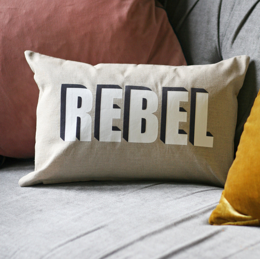 beige pillow with three dimensional white lettering reading 'rebel' and a black drop shadow 