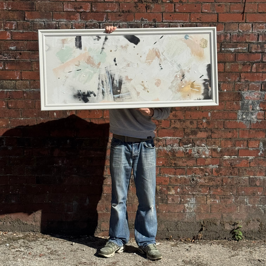 Person holding a framed abstract painting against a brick wall
