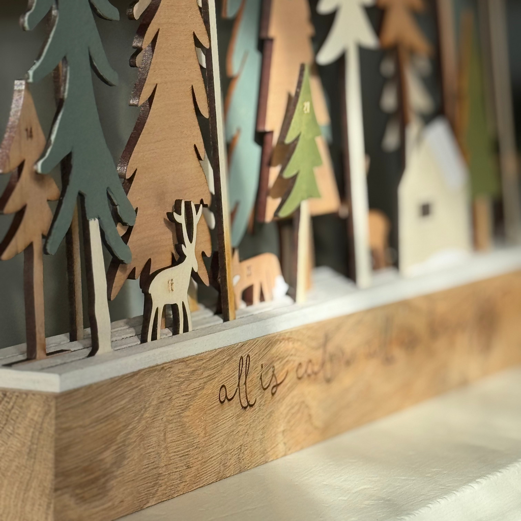 Wooden decorative scene with trees and a deer, featuring the text 'all is calm, all is bright'.