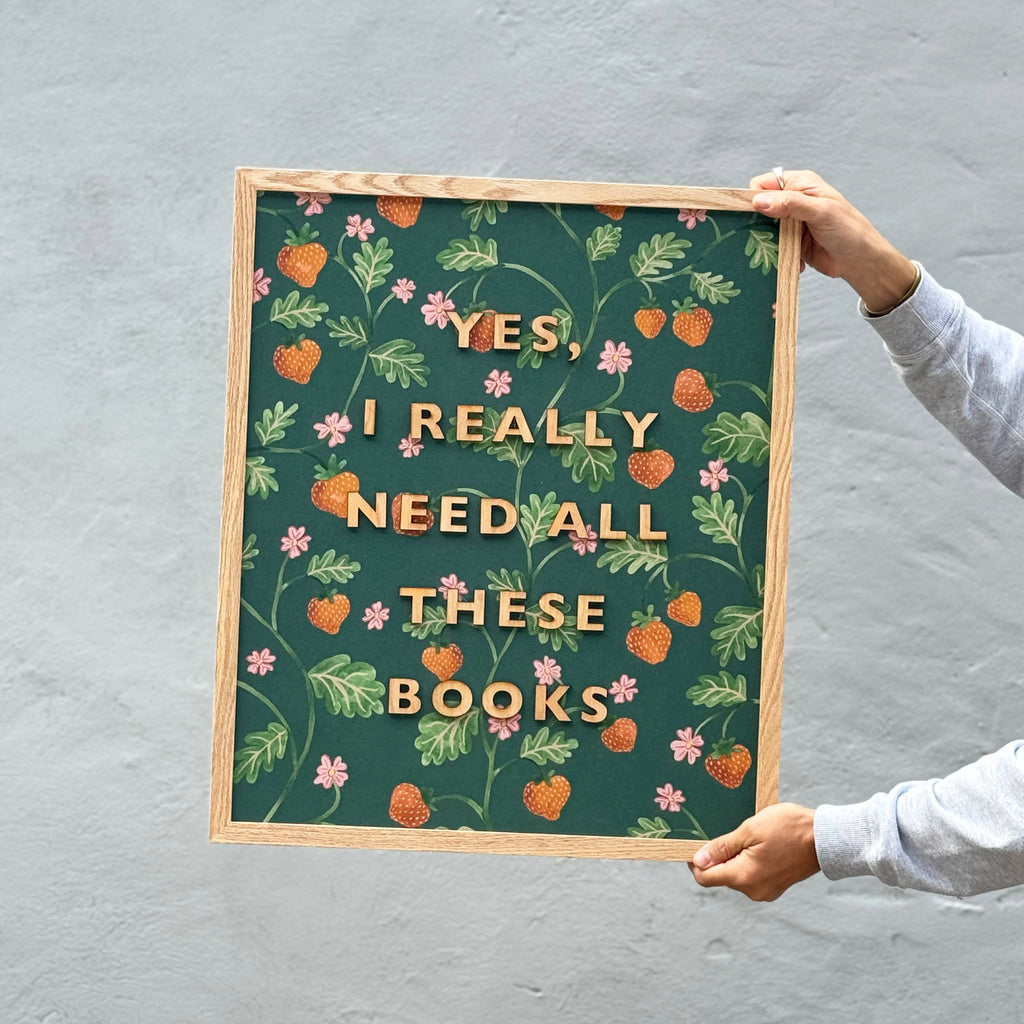 Person holding a framed print with 'Yes, I really need all these books' text and floral and strawberry pattern design on a gray background by modo