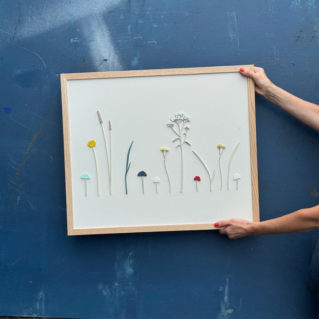 Framed hand painted artwork with floral and foliage designs held against a blue wall