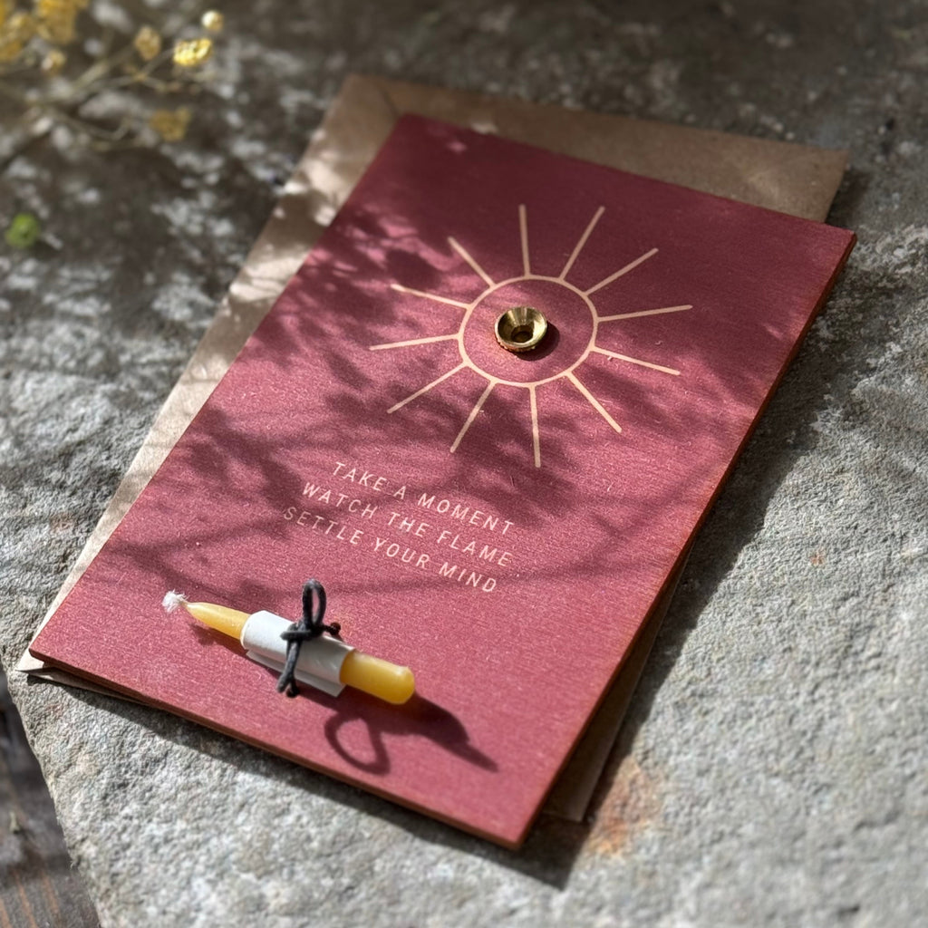 teracotta wooden mindful card with sun design , candle holder and beeswax candle and text on a stone surface with flowers.