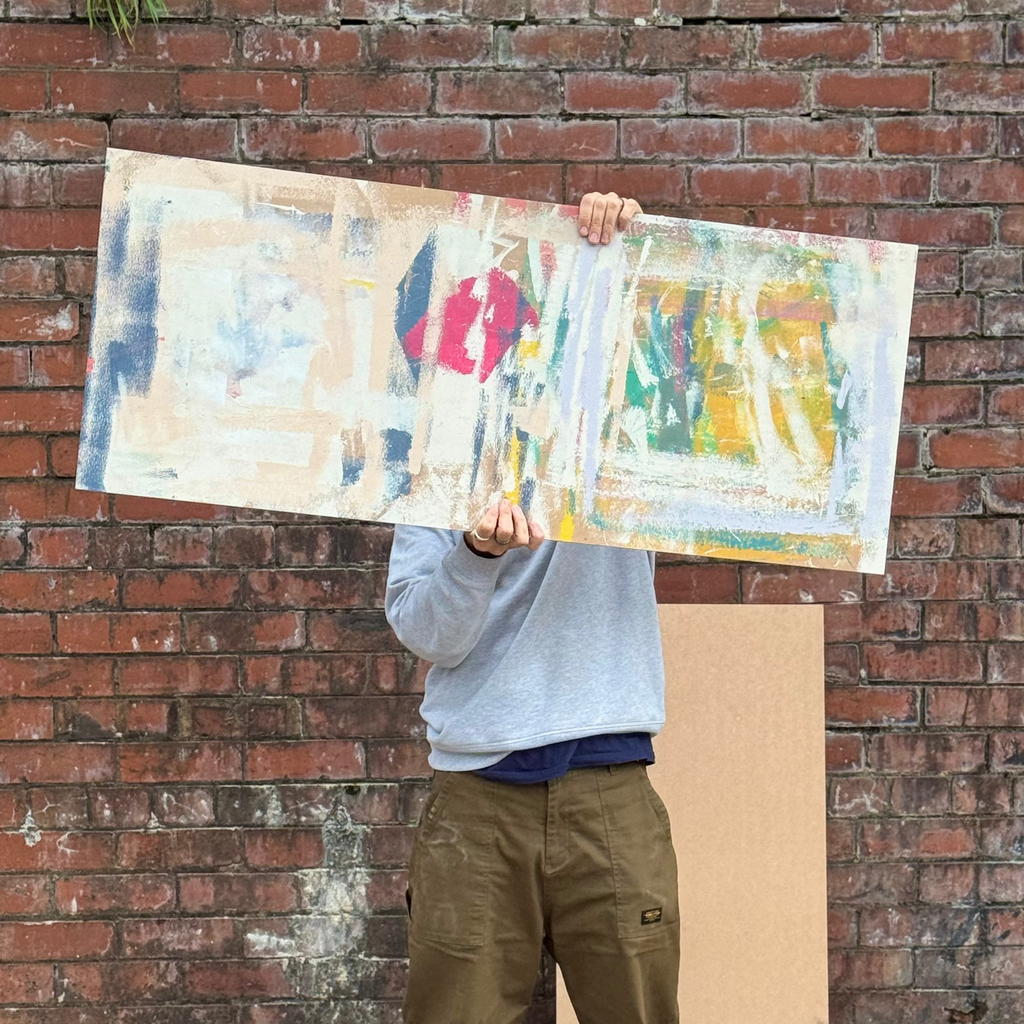Person holding a colorful abstract painting against a brick wall.