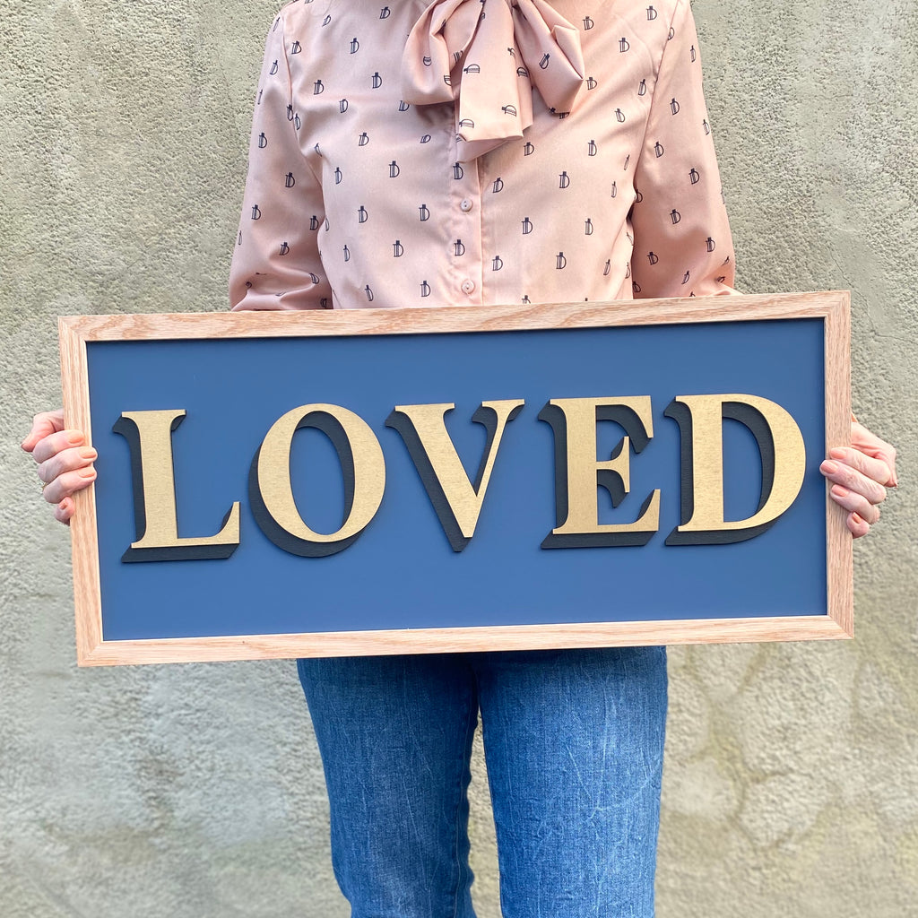 One word painted sign held by a person and displaying the word 'Loved' in gold