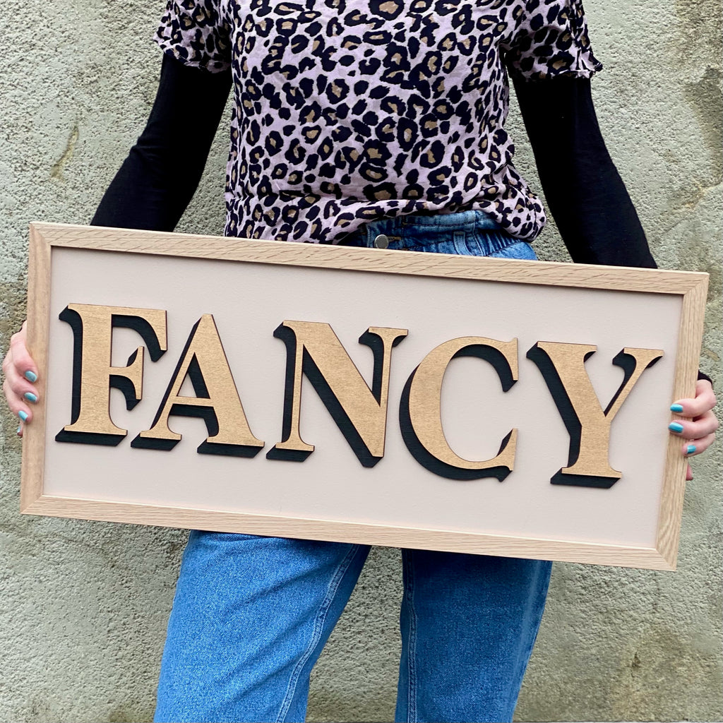 One word painted sign held by a person and displaying the word 'Fancy' in gold
