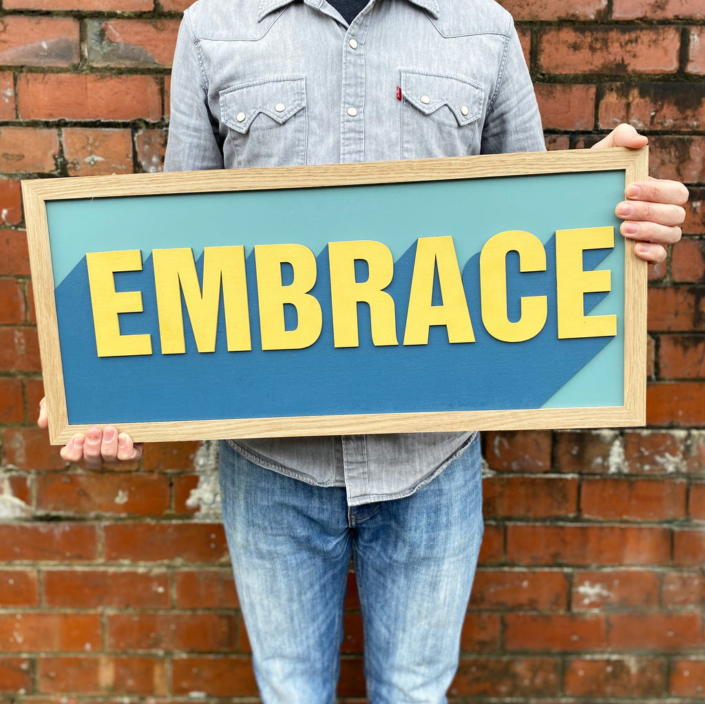 one word, hand painted layered sign displaying the word 'Embrace' in a yellow with contrasting blue and cyan backdrop.