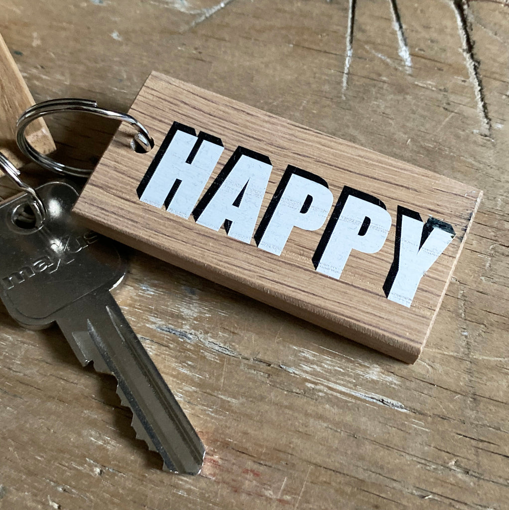 One word wooden keyring reading 'happy' in white box font with black shadow backdrop