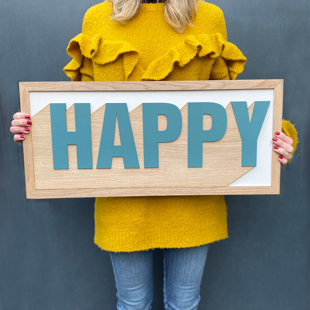 one word, layered, hand painted sign. the word 'happy' in blue with white and raw wooden background.
