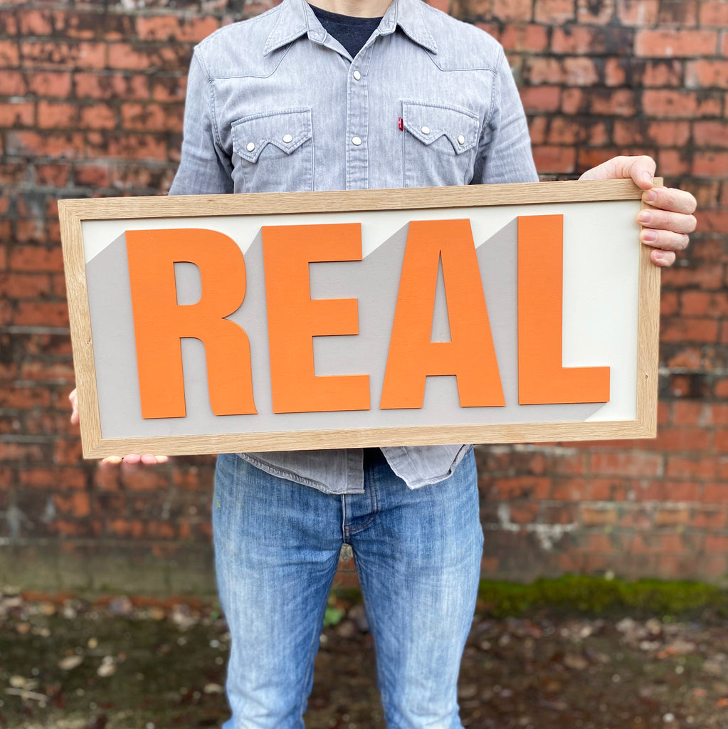One word layered hand painted sign displaying the word 'real' in a vibrant orange colour with white and grey backdrop