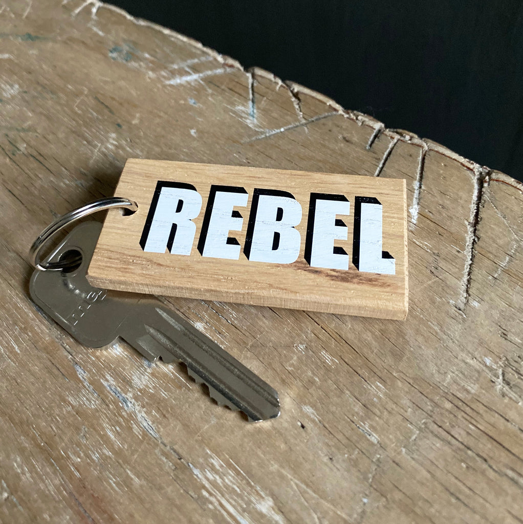 One word wooden keyring reading 'rebel' in white box font with black drop shadow